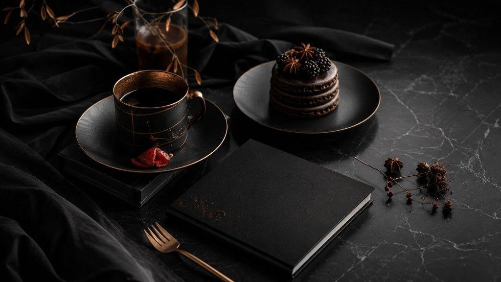 Dark noir still life on black marble: matte black book, copper-rimmed coffee cup, stacked dark chocolate desserts with blackberries and star anise, and a tall glass of dark beverage.