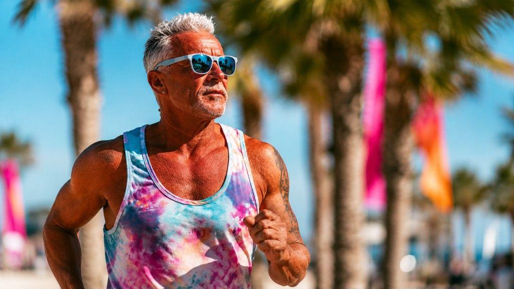 Athletic man jogging on a sunlit seaside path in a bright tie-dye tank top and blue sunglasses, with palm trees and colourful summer banners behind him.